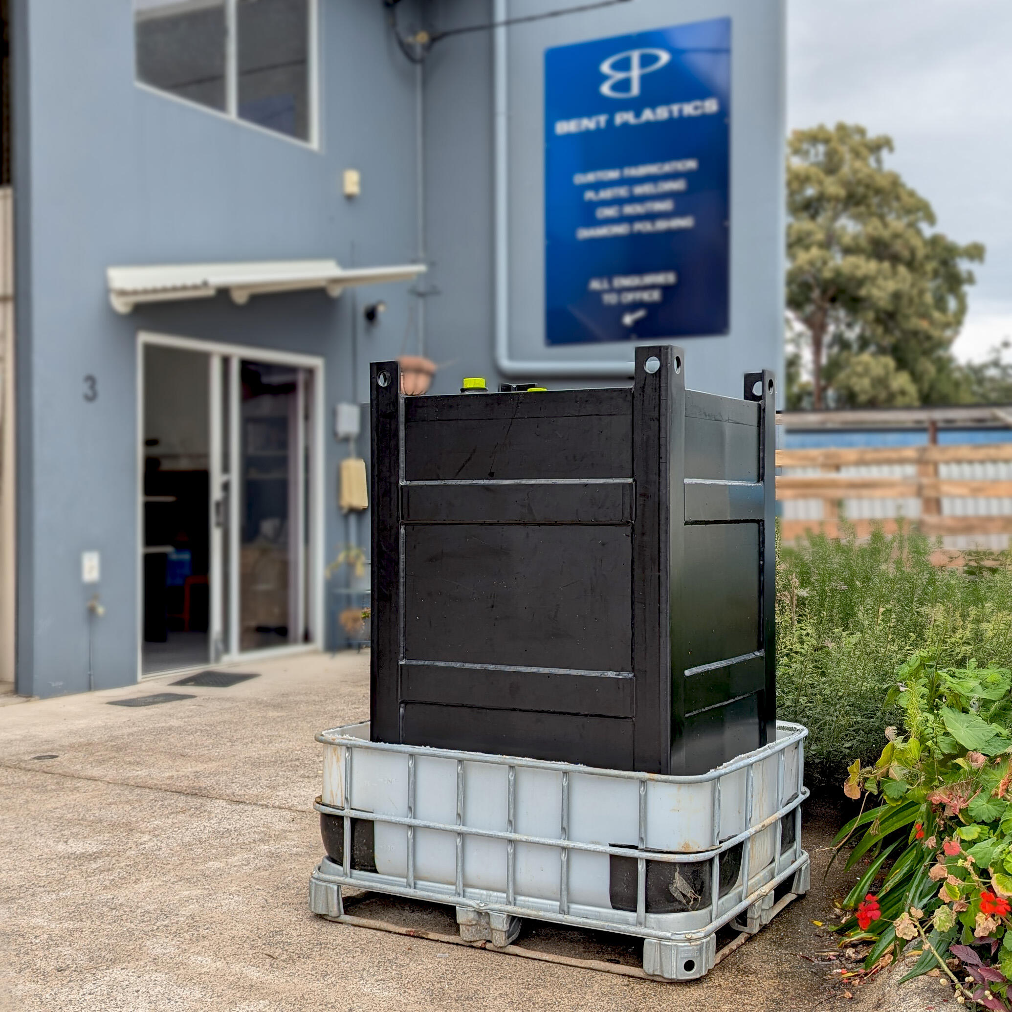 Rectangular black plastic tank sitting in a metal and plastic tray, in front of a factory with a sign that says Bent Plastics